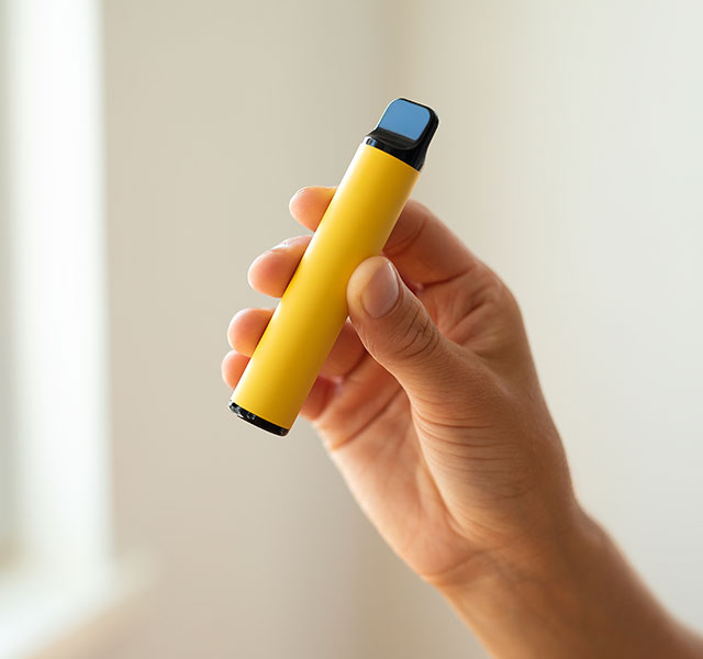 A hand holding a yellow vape pen near a window with soft light filtering in. fruit flavored ecigs