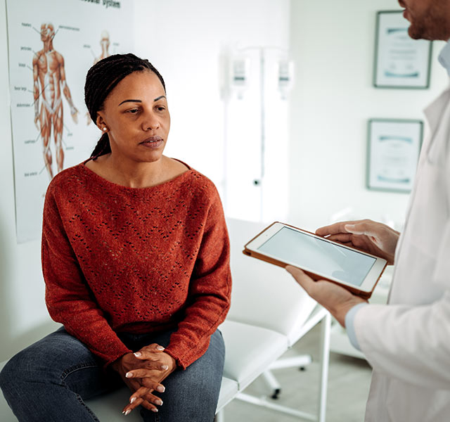 A healthcare professional holding a tablet, speaking with a woman sitting on an examination table. There is a medical diagram on the wall in the background, suggesting a clinical setting. pmos