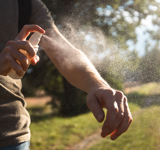 A person outdoors sprays insect repellent onto their arm, with sunlight filtering through trees in the background. repelling mosquitoes
