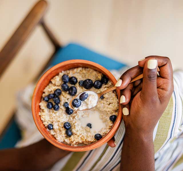 A person holds a bowl of oatmeal topped with blueberries and milk, using a spoon. dummy mobile image
