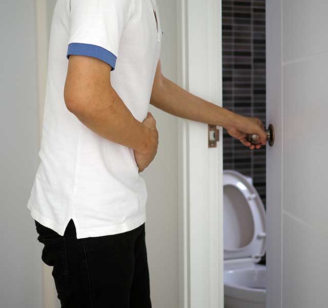 A person wearing a white shirt with blue trim is holding their stomach while opening a door to a bathroom. A toilet with an open lid is visible in the background. chronic constipation