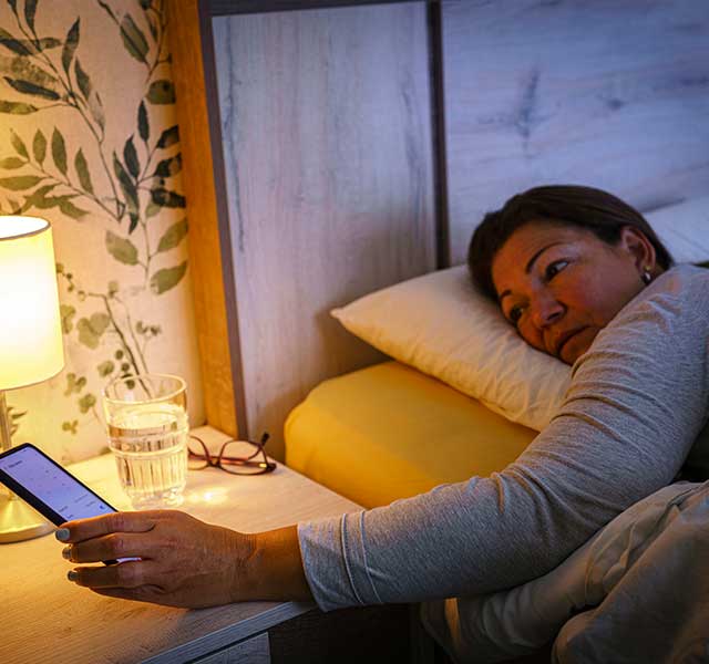 A person lies in bed, reaching towards a smartphone on a bedside table. A lamp provides soft lighting, and a glass of water and a pair of glasses are also on the table. diabetes and sleep