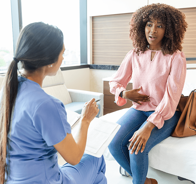 Young woman with brown curly hair in a pink long-sleeve shirt and jeans sitting in the doctor's office talking to doctor. stomach noises