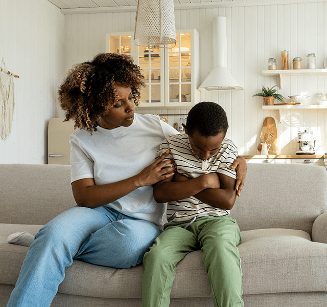 Mom with brown curly short hair wearing a white T-shirt and jeans. She is sitting on a couch trying to comfort her young son who is wearing a white and navy blue striped t-shirt and green pants. He has his arms folded over his chest. dummy mobile image