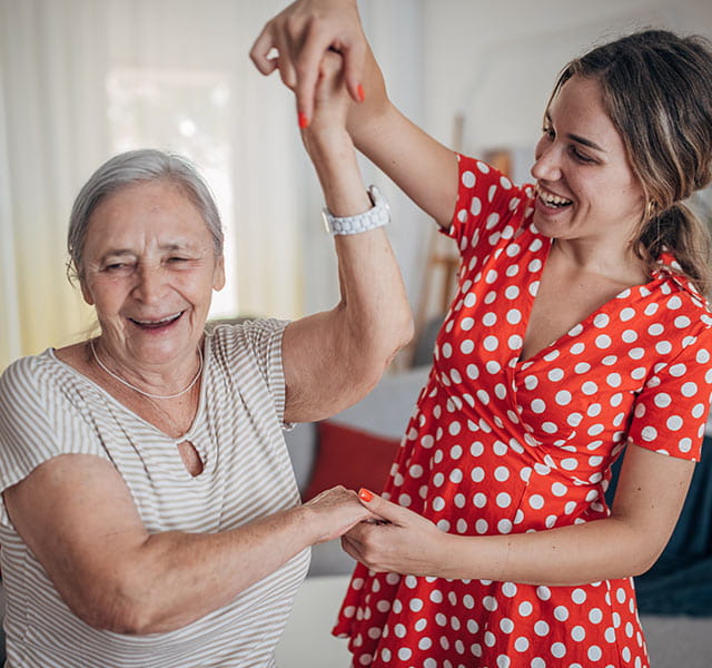 Two women are smiling and dancing indoors. One person is older with grey hair and the other is younger wearing a red polka dot top. The setting appears to be a bright room with curtains in the background. everyday exercises for brain health