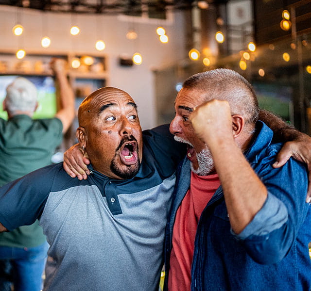 A group of people excitedly celebrating in a room, likely due to a sports event on a TV in the background. Two people in the foreground are embracing with joyful expressions, surrounded by warm lighting and hanging bulbs. sporting event heart attack