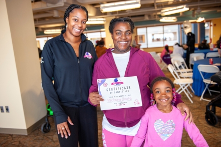 A WIN Network mother stands with her daughter and her community health worker while holding her BLOSSOM Program Certificate of Completion, recognizing her participation and achievement.